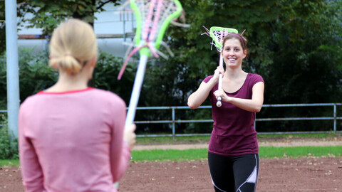 Zwei Personen in Sportkleidung stehen sich auf einem Sportplatz gegenüber und spielen Lacrosse.