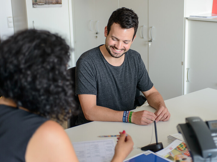 Zwei Peronen sitzen an einem Tisch und führen ein Beratungsgespräch. Student lächelt, Beraterin zeigt mit einem Stift auf ein Blatt Papier, auf dem ein Stundenplan zu sehen ist.