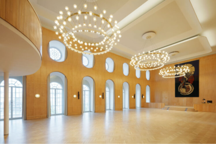 Aula der Uni Mannheim mit hölzernen Wänden, hohen Fenstern, drei runden Kronleuchtern und einem Podium.