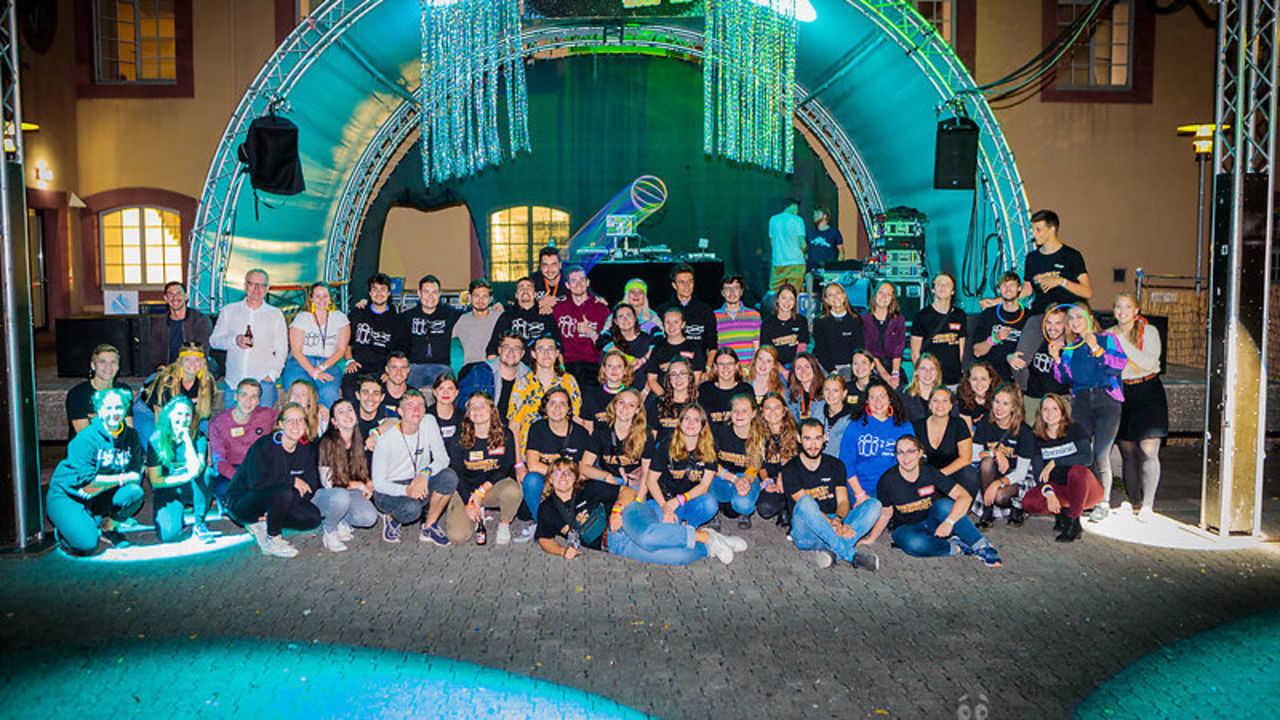 Ein Gruppenfoto von über 50 lächelnden Studierenden während einer Party. Im Hintergrund steht eine beleuchtete Bühne.