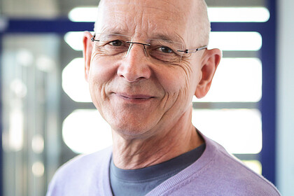 Portrait von Rüdiger Schmitt-Beck. Er trägt einen lila farbenen Pullover und steht lächelnd vor einer Glastüre.