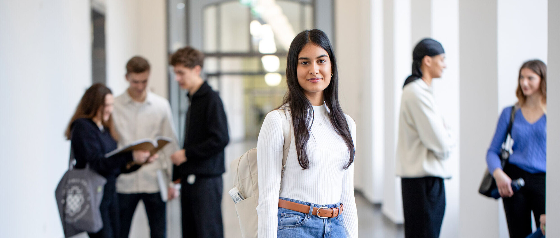 Eine Studenin steht im Flur des Schlosses und schaut direkt und offen in die Kamera. In der Hand hält sie ein Buch. Im Hintergrund stehen fünf Studierenden unterhalten sich. Sie haben Laptops, Taschen udn Ruchsäcke dabei.