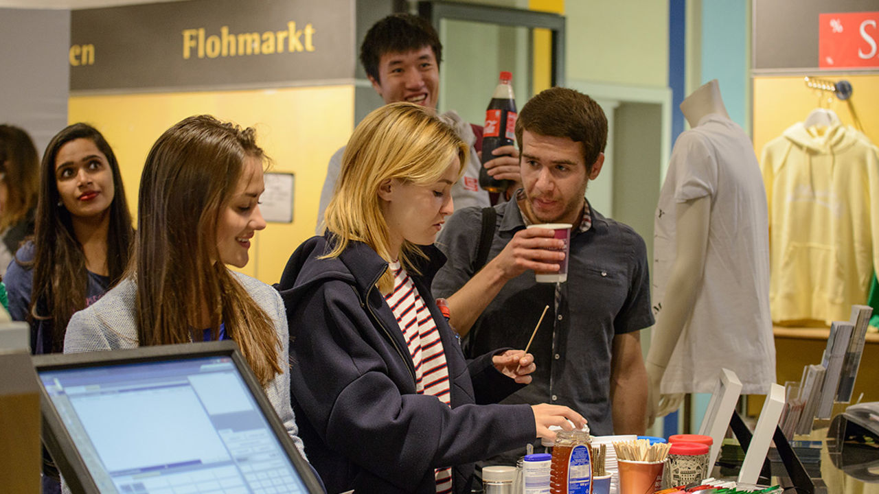Studierende stehen gutgelaunt vor dem Campus Shop der Universität Mannheim. Vor ihnen steht ein Tablet mit Zutaten für Kaffee und Tee.