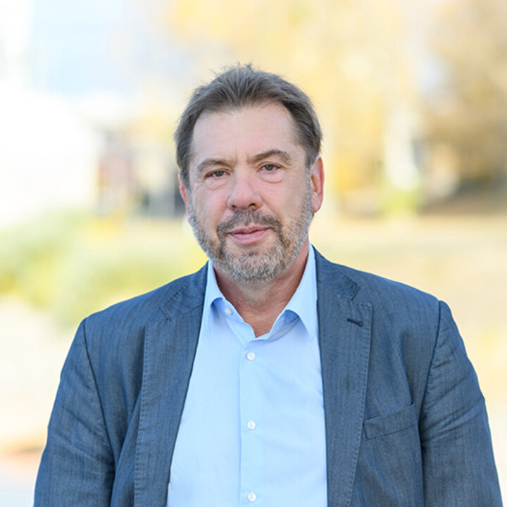 Georg Licht draußen vor einem grünen Hintergrund. Er trägt ein hellblaues Hemd unter einem dunklen Blazer.