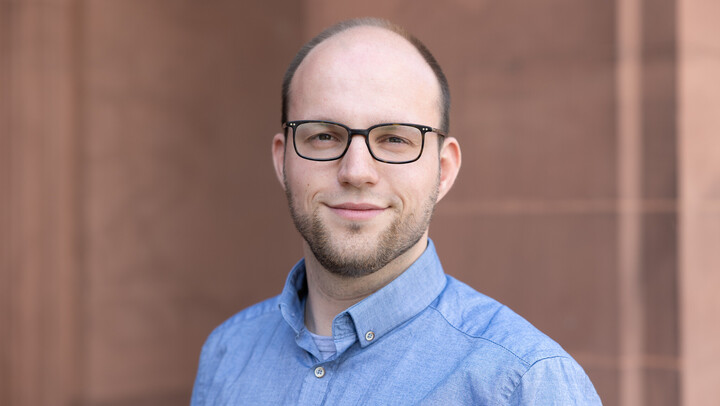 Malte Grönemann. He is wearing a blue shirt and is standing in front a brown stonewall.