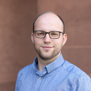 Portrait of Malte Grönemann. He is wearing a blue shirt and standing next to a building.