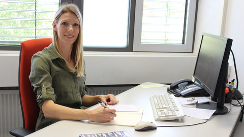 Eine Person trägt ein olive-farbenes Kleid und sitzt an einem Schreibtisch. Die Person heißt Johanna Nickolai.