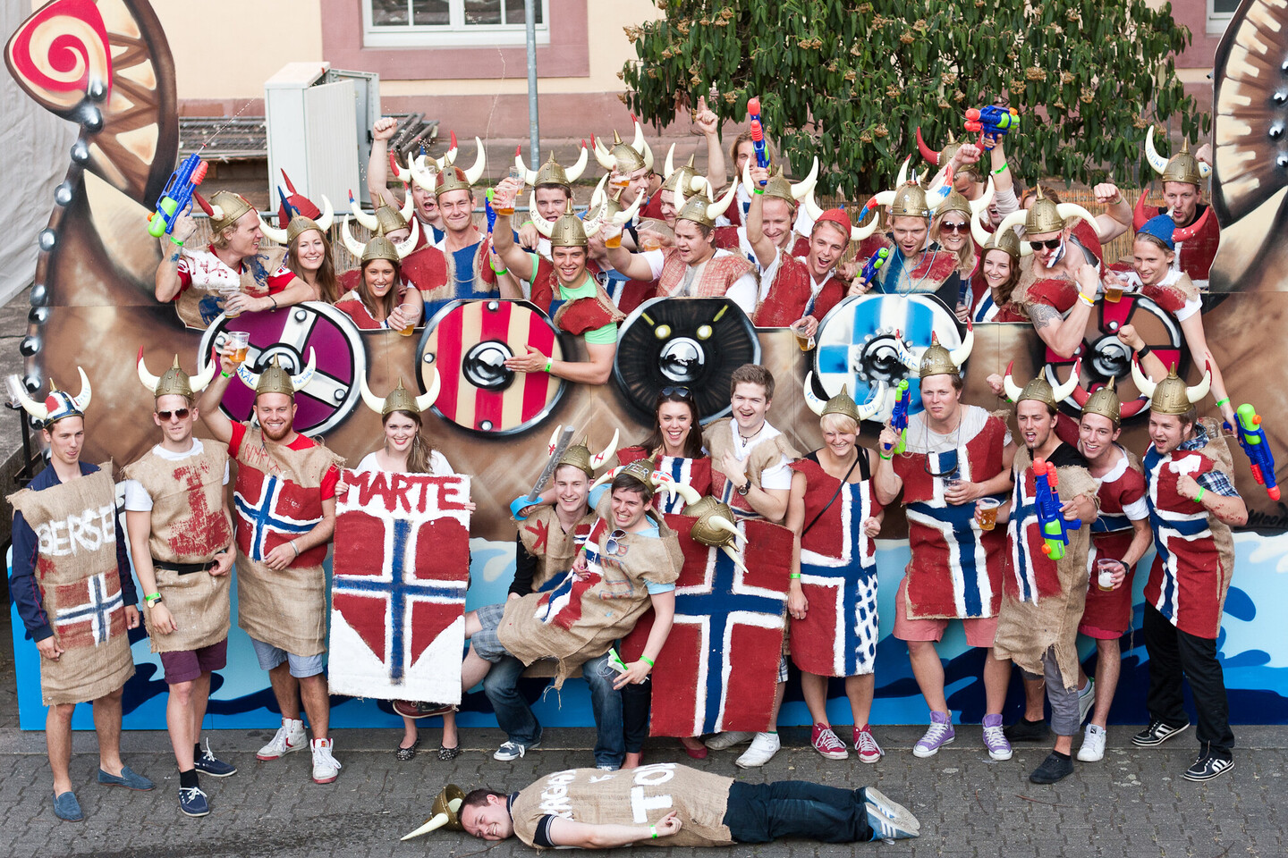 Etwa 30-40 Menschen, die als Wikinger verkleidet sind, stehen vor einem aus Pappe gebastelten Wikingerboot. Sie tragen Wikingerhelme mit zwei Hörnern und viele die norwegische Flagge als Gewand.