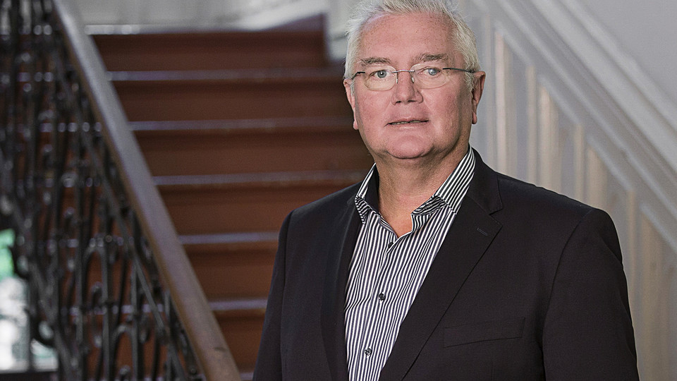 Prof. Homburg hat graue Haare und eine Brille. Er trägt Hemd und Jacket und steht vor einem Treppenaufgang.
