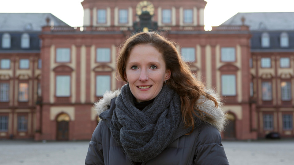 Portrait von Karin Hoisl. Sie trägt eine schwarze Jacke und einen grauen Schal, im Hintergrund ist das Mannheimer Schloß. 