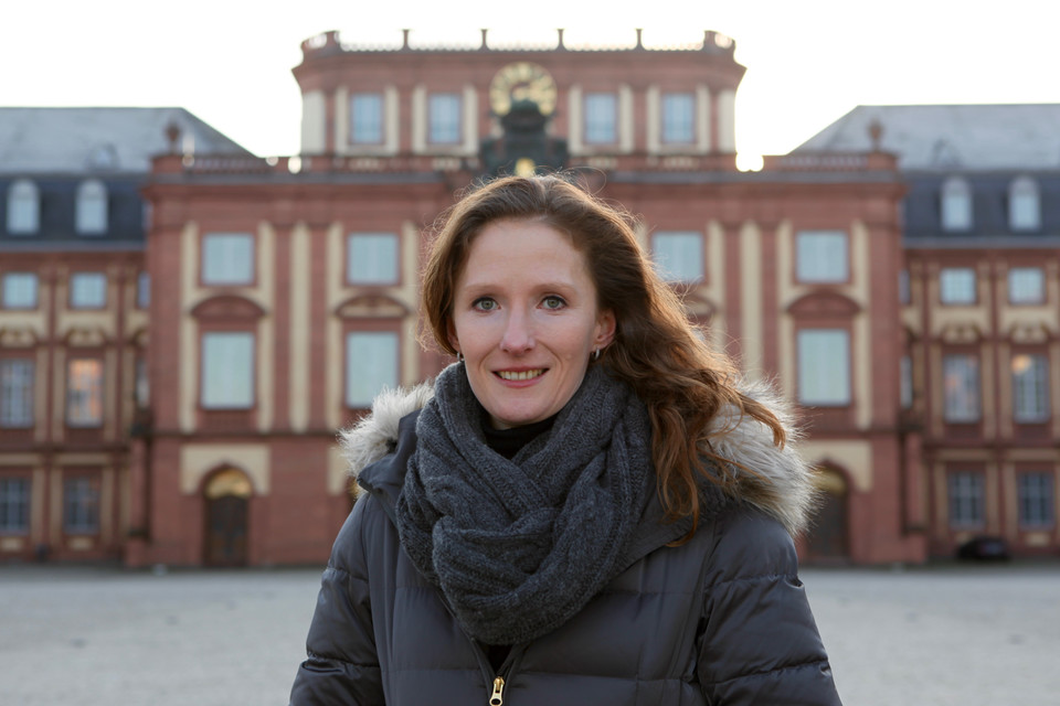 Portrait von Karin Hoisl. Sie trägt eine schwarze Jacke und einen grauen Schal, im Hintergrund ist das Mannheimer Schloß. 