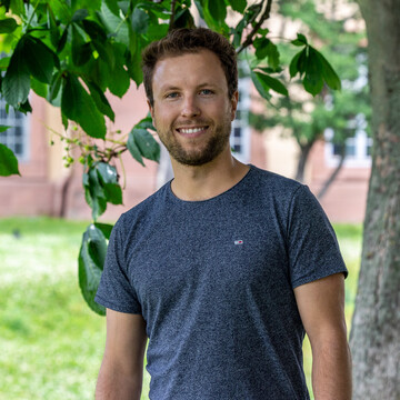Philip Holler trägt ein dunkelblaues T-Shirt und steht vor einem Baum auf der Mensawiese. Er lächelt in die Kamera.