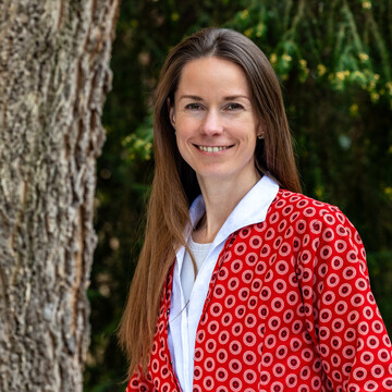 Prof. Dr. Ilze Plavgo. Sie steht lächelnd vor einem Baum und trägt einen rot gemusterten Blazer.
