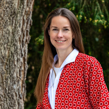 Prof. Dr. Ilze Plavgo. Sie steht lächelnd vor einem Baum und trägt einen rot gemusterten Blazer.