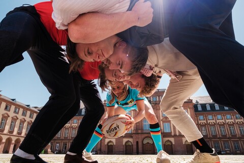 Am Ehrenhof stehen vier Rugbyspieler in formeller Kleidung in Startposition. Eine Rugbyspielerin in blauer Uniform steht hinter ihnen in vorgebeugter Position mit einem Ball in der Hand.