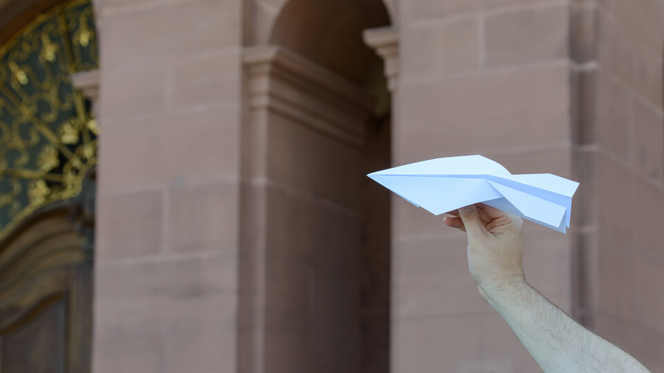 Eine Hand hält einen Papierflieger vor der Kulisse des Mannheimer Schlosses im Hintergrund.