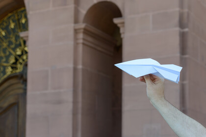 Eine Hand hält einen Papierflieger vor der Kulisse des Mannheimer Schlosses im Hintergrund.