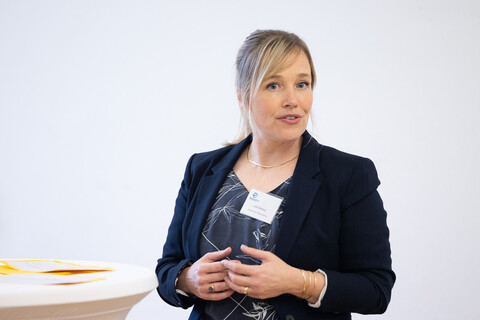Julia Derkau trägt einen dunkelblauen Blazer sowie eine blau gemusterte Bluse, steht neben einem Stehtisch und spricht.