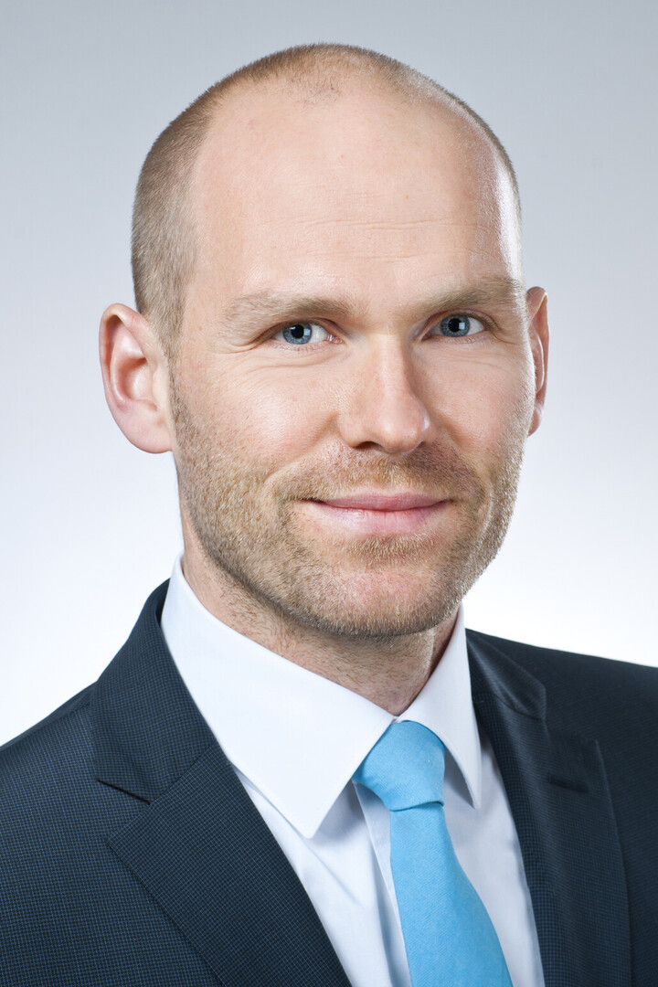 Eine Person trägt ein weißes Hemd, eine blaue Krawatte sowie einen dunkelblauen Blazer und steht vor einem weißen Hintergrund. Die Person heißt Florian Kutzner.