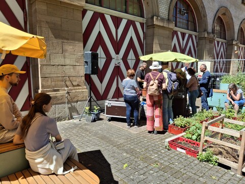 Mann mit Gitarre, mehrere sitzende Leute die zum Mann mit Gitarre schauen vor einem Haus mit rot-weiß-gestreiften Holztüren - die Sonne scheint