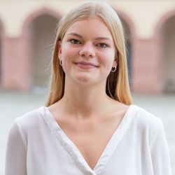 Anna Lenz hat blonde lange Haare. Sie trägt Creolen-Ohrringe und ein weißes Hemd und steht auf dem Ehrenhof vor dem Mannheimer Schloss.