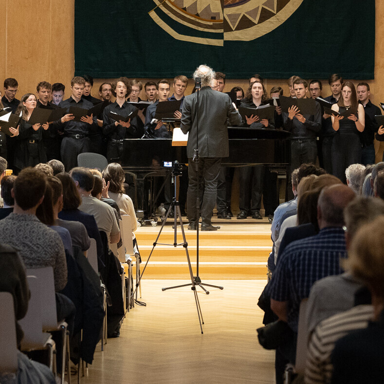 Auf der Bühne steht der Universitätschor mit Dirigent, im Vordergrund sieht man das Publikum, das zuhört.