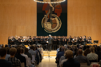 Auf der Bühne steht der Universitätschor mit Dirigent, im Vordergrund sieht man das Publikum, das zuhört.