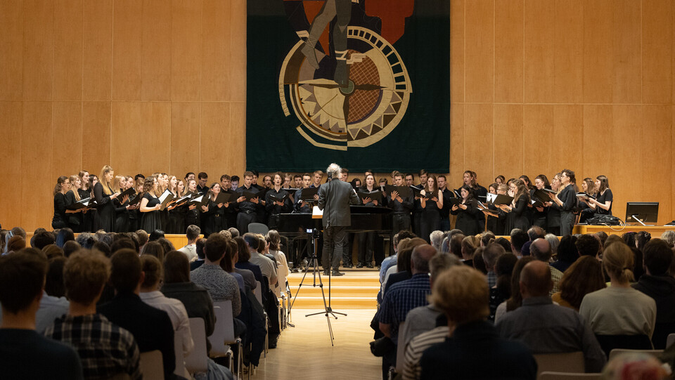 Auf der Bühne steht der Universitätschor mit Dirigent, im Vordergrund sieht man das Publikum, das zuhört.