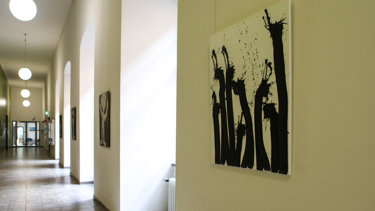 Ein Flur mit Bögen, welche an der rechten Wand entlanglaufen, ist geschmückt mit Kunstwerken. Abgebildet sind schwarze Farbklecks-Figuren und Zeitungscollagen.