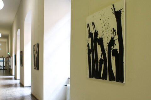 Ein Flur mit Bögen, welche an der rechten Wand entlanglaufen, ist geschmückt mit Kunstwerken. Abgebildet sind schwarze Farbklecks-Figuren und Zeitungscollagen.