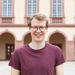 Ein Porträt von Konstantin Werner. Er hat kurzes blondes Haar und trägt eine braune runde Brille sowie ein dunkelrotes T-Shirt.