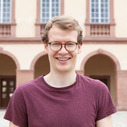 Konstantin Werner hat kurzes blondes Haar. Er trägt eine braune runde Brille und ein dunkelrotes T-Shirt.