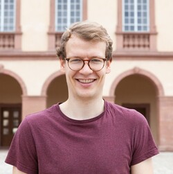 A portrait of Konstantin Werner. He has short blond hair and is wearing round brown glasses and a dark red T-shirt.