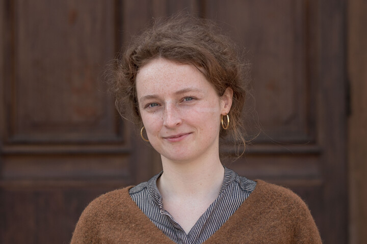 Ursula Berresheim. Sie trägt einen hellbraunen Pullover und steht vor dunkelbraunen Holztür.