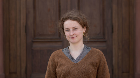 Ursula Berresheim. She is wearing a brown sweater and is standing in front of a brown door.