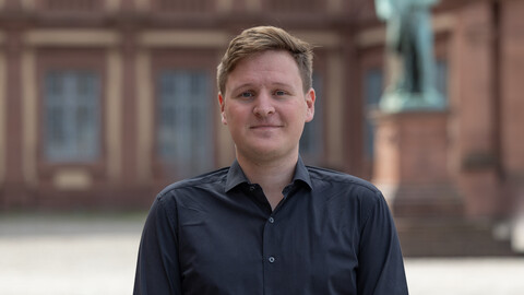 Janko Wilzbach. He is wearing a black shirt and is standing on the Ehrenhof in front of the castle.