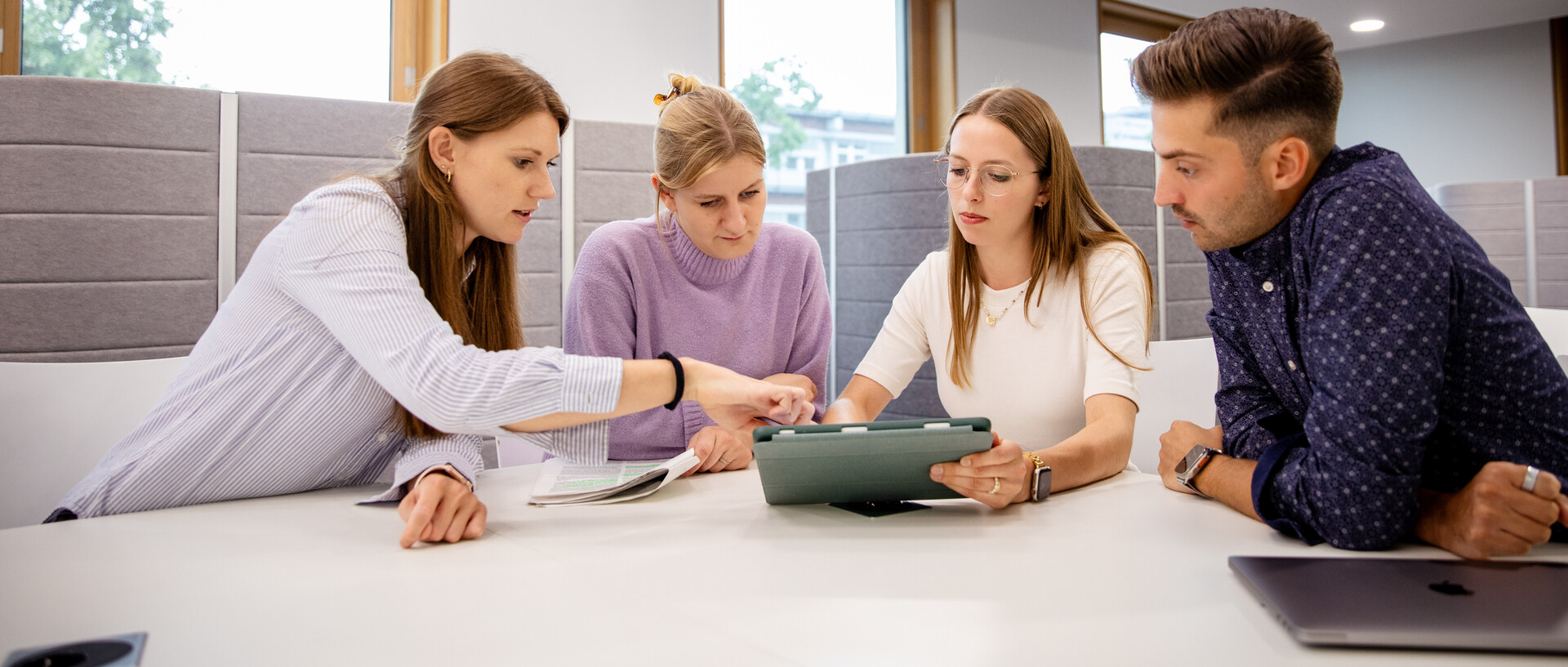 Vier Personen sitzen an einem Tisch und blicken auf ein Tablet.