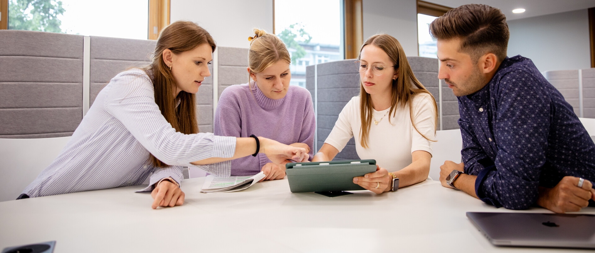 Vier Personen sitzen an einem Tisch und blicken auf ein Tablet.
