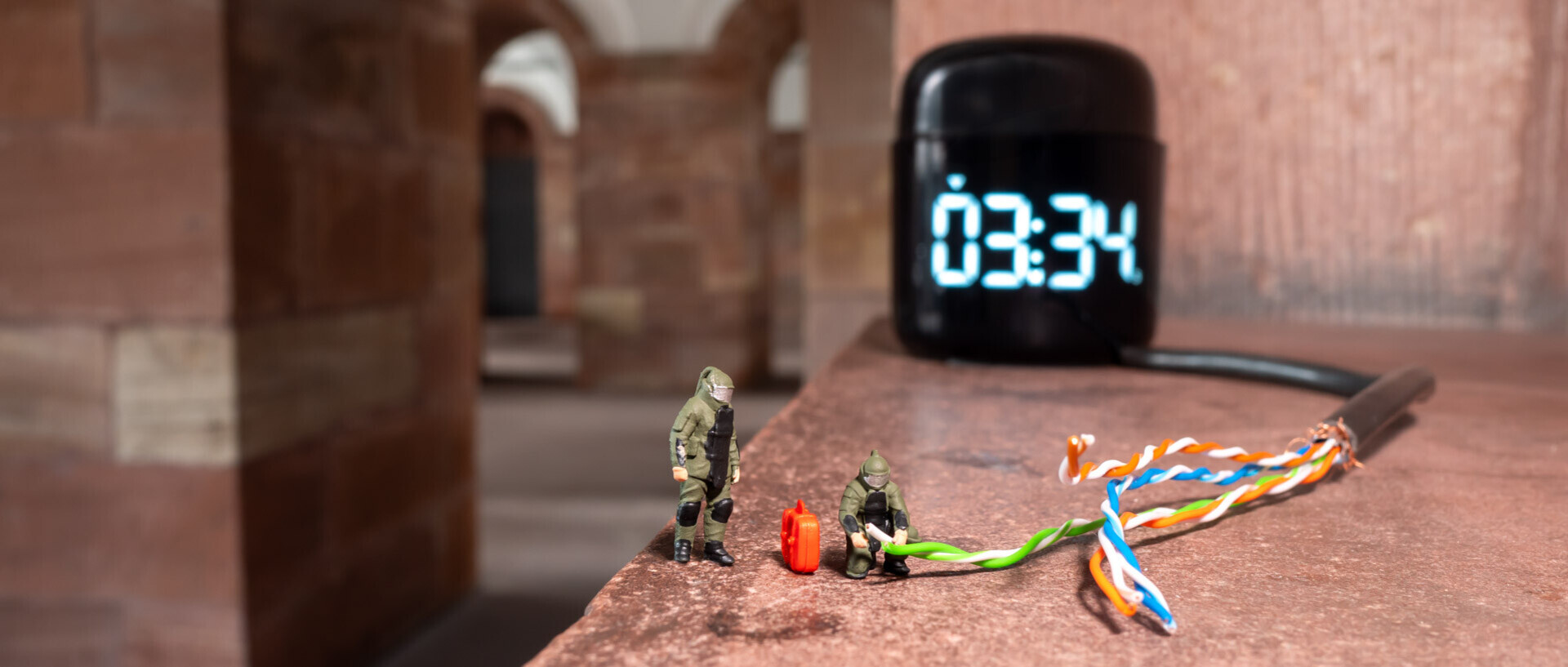In the photo, two miniature figures are defusing a bomb, which is represented by a timer and different colored cables.