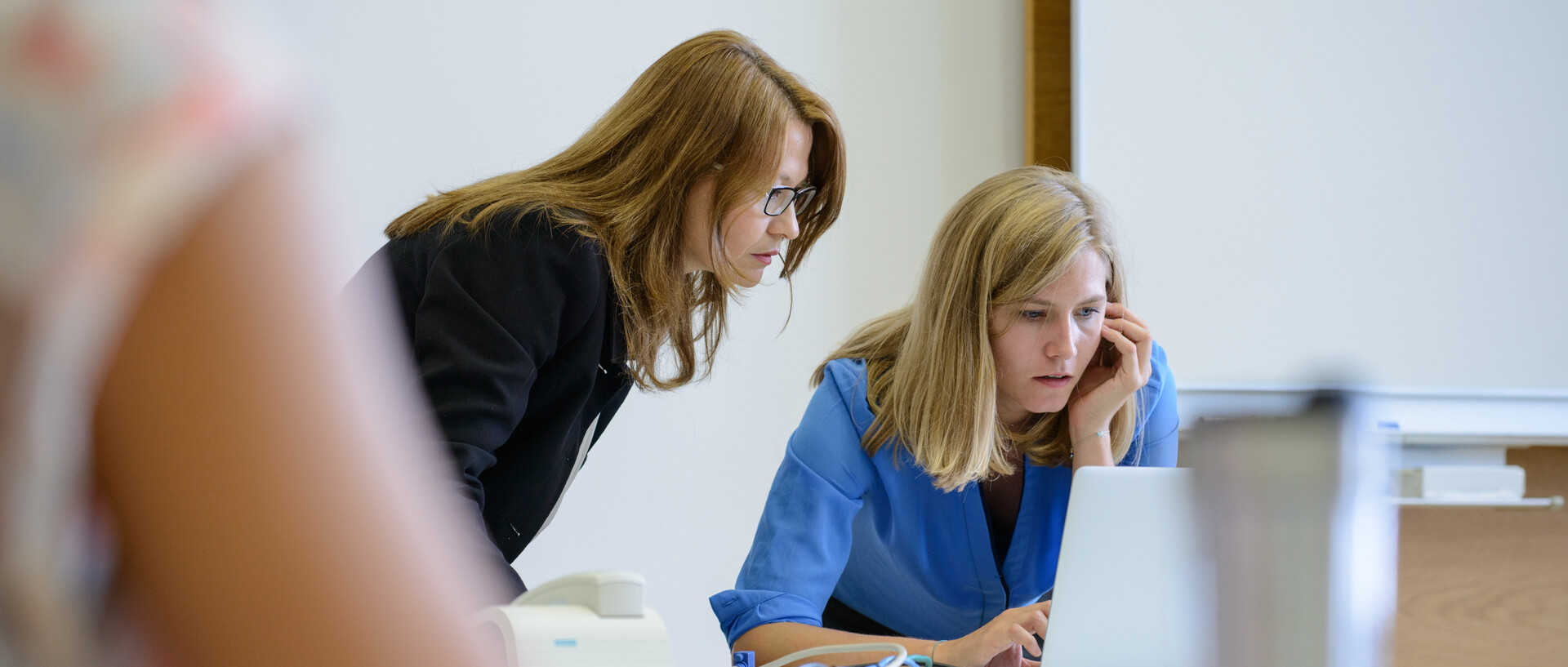 Zwei Frauen schauen zusammen auf den Display eines Laptops. Sie stehen vorne an der Tafel.