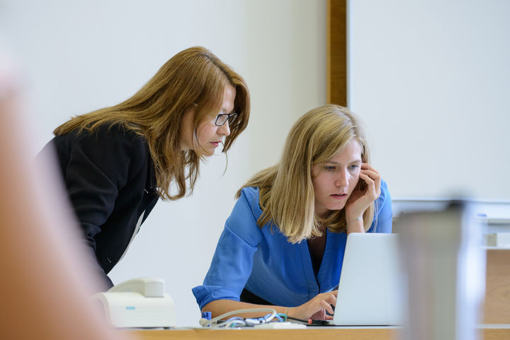 Zwei Frauen schauen zusammen auf den Display eines Laptops. Sie stehen vorne an der Tafel.