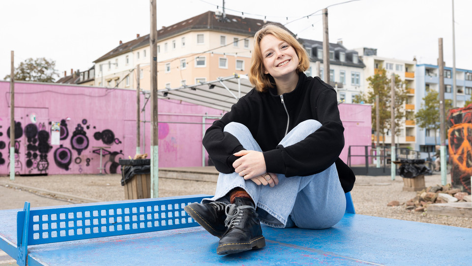 Eine Studentin mit kurzen blonden Haaren, einem schwarzen Pulli und einer hellen Jeans sitzt auf einer blauen Tischtennisplatte.