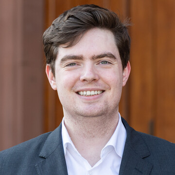 Portrait von Tim Hoeckmann. Er trägt einen Anzug mit weißem Hemd und steht vor einer brauen Tür der Universität.