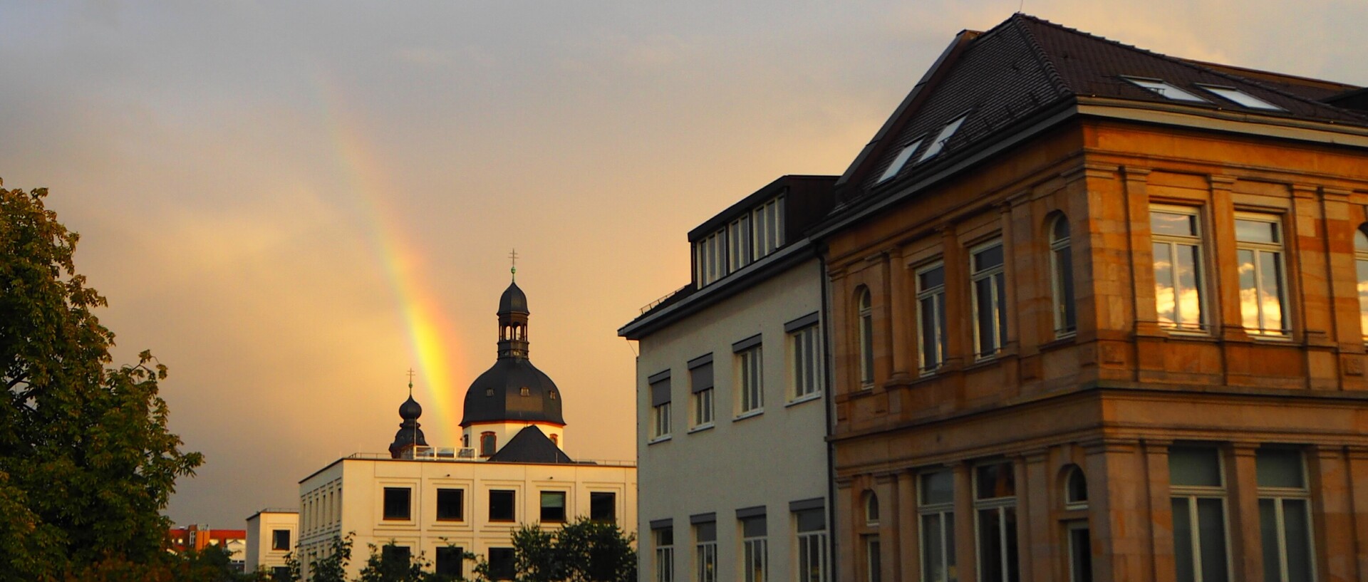 Gebäude werden von der Abendsonne beleuchtet. Im Hintergrund ist ein Kirchturm mit einem Regenbogen zu sehen.