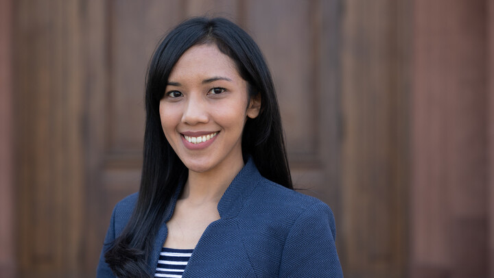Sandra Kurniawati. She is wearing a blue-white stripped shirt and a blue jacket and is standing in front of a brown door.