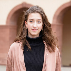 Pamina Heermann hat braune lange Haare und trägt Creolen-Ohrringe. Sie trägt einen schwarzen Strickrollkragenpullover und darüber ein lockeres rostbraunes Hemd. Sie steht vor dem Mannheimer Schloss. 