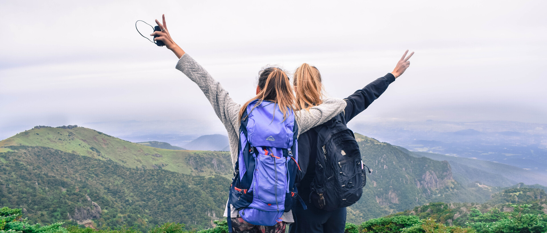 Im Vordergrund stehen zwei junge Frauen, die sportlich gekleidet sind, große Rucksäcke tragen und ihre Arme in die Luft strecken. Sie blicken in die Ferne. Der Hintergrund zeigt eine Panoramalandschaft mit vielen grünen Hügeln.