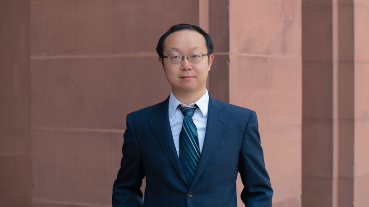 Chen Lin. He is wearing a dark blue suit and is standing in front of a brown stonewall.