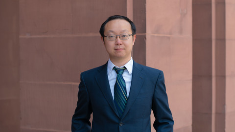 Chen Lin. He is wearing a dark blue suit and is standing in front of a brown stonewall.