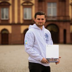 Ein Stipendiat des Spitzensport-Stipendiums steht vor dem Mannheimer Schloss und trägt einen grauen Hoodie der Universität mit verschiedenen Logos. Er hält eine Mappe der Universität Mannheim in seinen Händen.